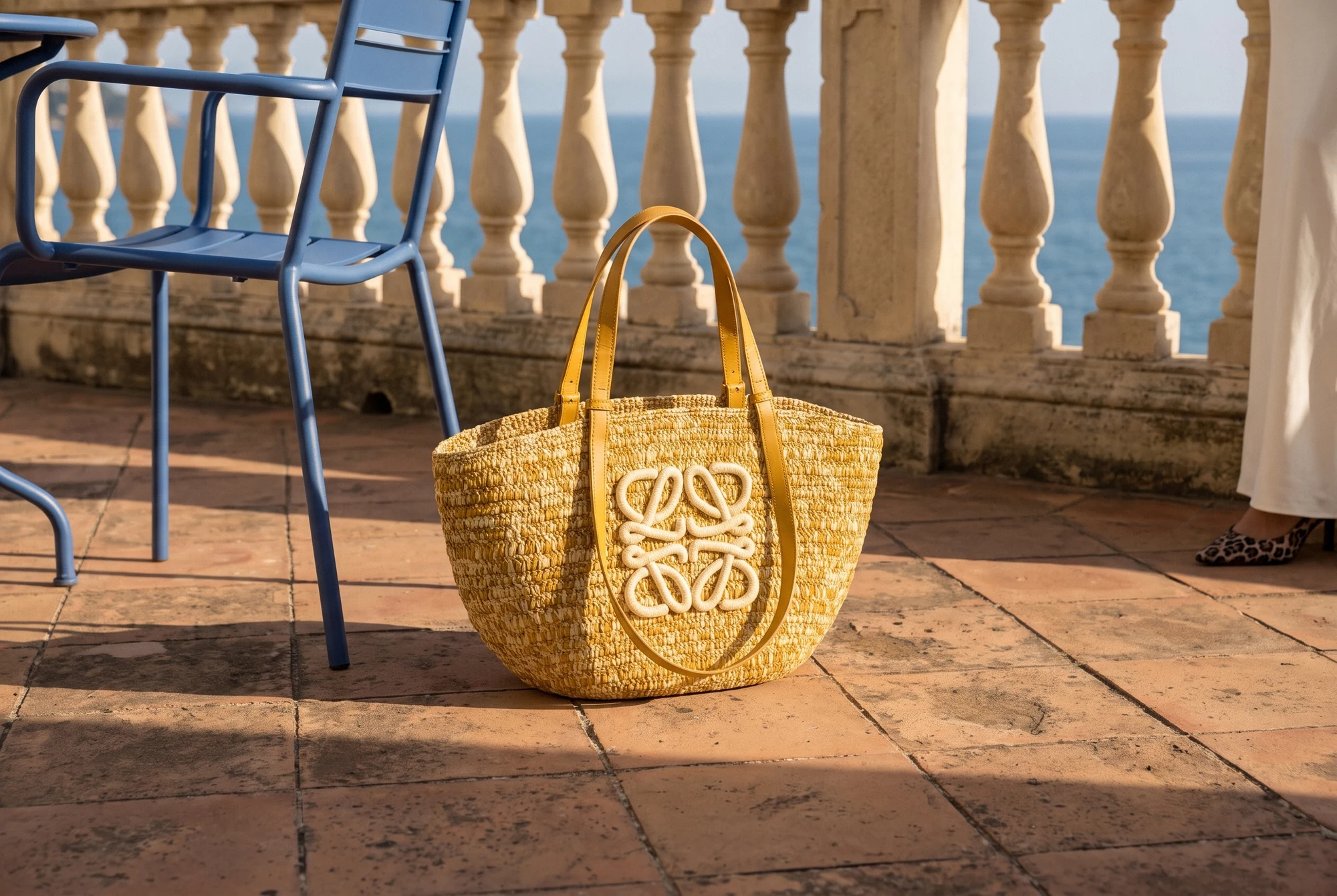 Bolso de rafia Loewe con anagrama junto a una silla azul y la balaustrada